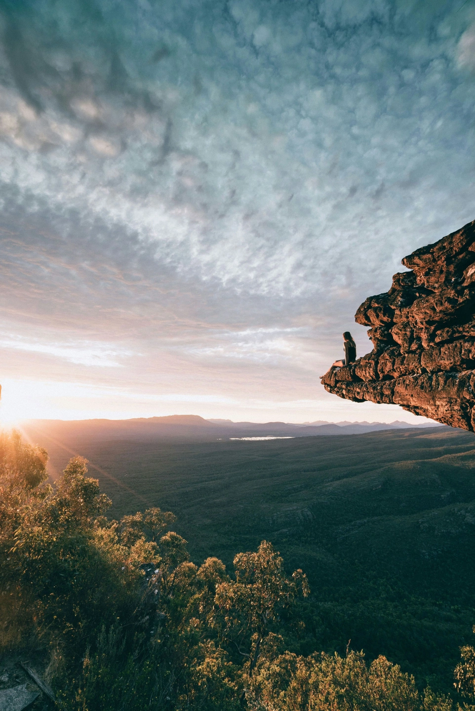 A person seated on a rock ledge overlooking a forested valley at sunrise